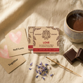 LOVE Ritual Set met Amma Cacao bar, ritueelkaarten voor Self Love en Together, blauwe korenbloemblaadjes en wierook op een beige linnen achtergrond.