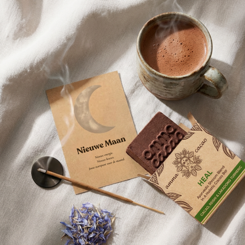 Nieuwe Maan cacao ritueel met Heal Amma Cacao bar, Nieuwe Maan intentiekaart met maansikkel, wierookstokje en blauwe bloemblaadjes.